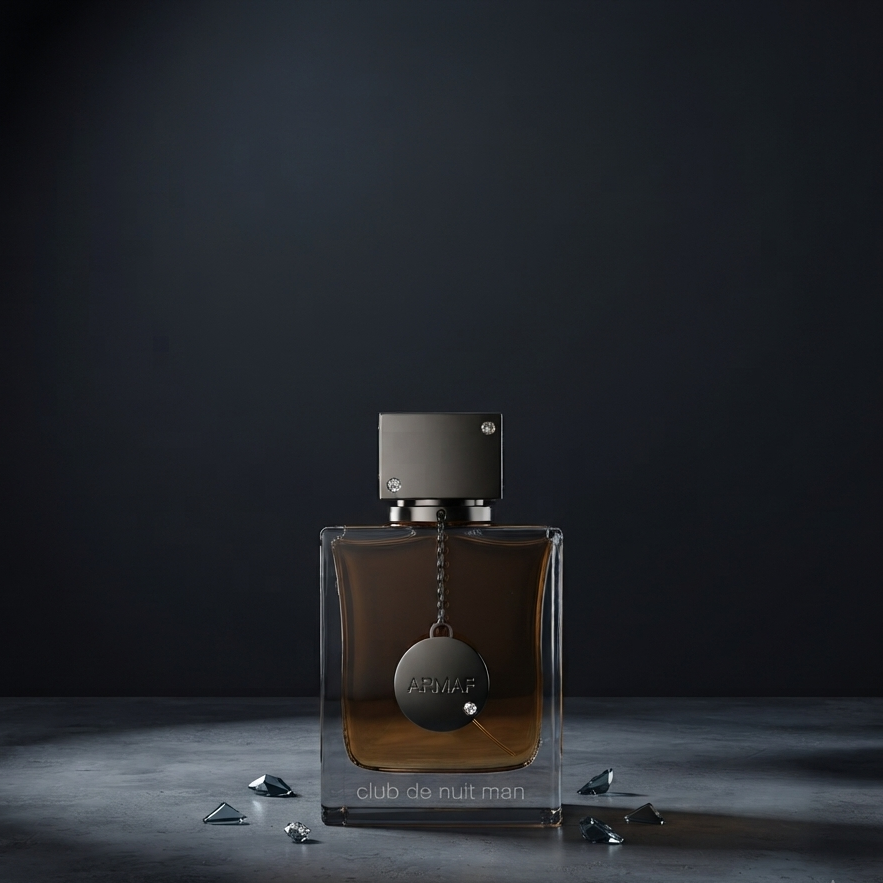 A rectangular clear glass perfume bottle with amber-brown liquid, a dark metallic cap, and a hanging round medallion, displayed on a dark surface against a dark background. Small faceted crystal-like pieces are scattered around the base. SHE GLOW