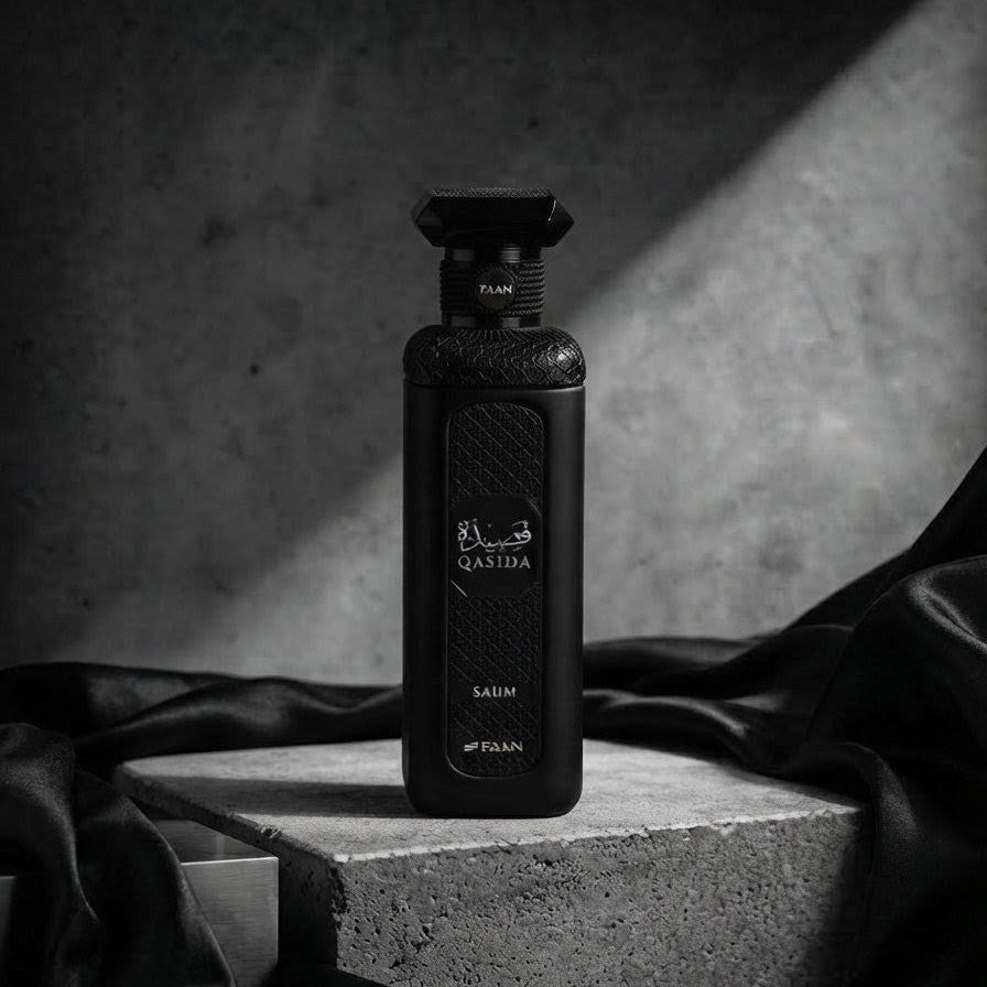 A black rectangular perfume bottle labeled 'FLAME' and 'SAUM' stands on a concrete pedestal with draped black fabric around it against a dark textured gray background. SHE GLOW ✨