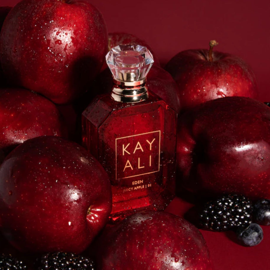 Red perfume bottle with 'KAY ALI' branding surrounded by red apples and black berries on a red background