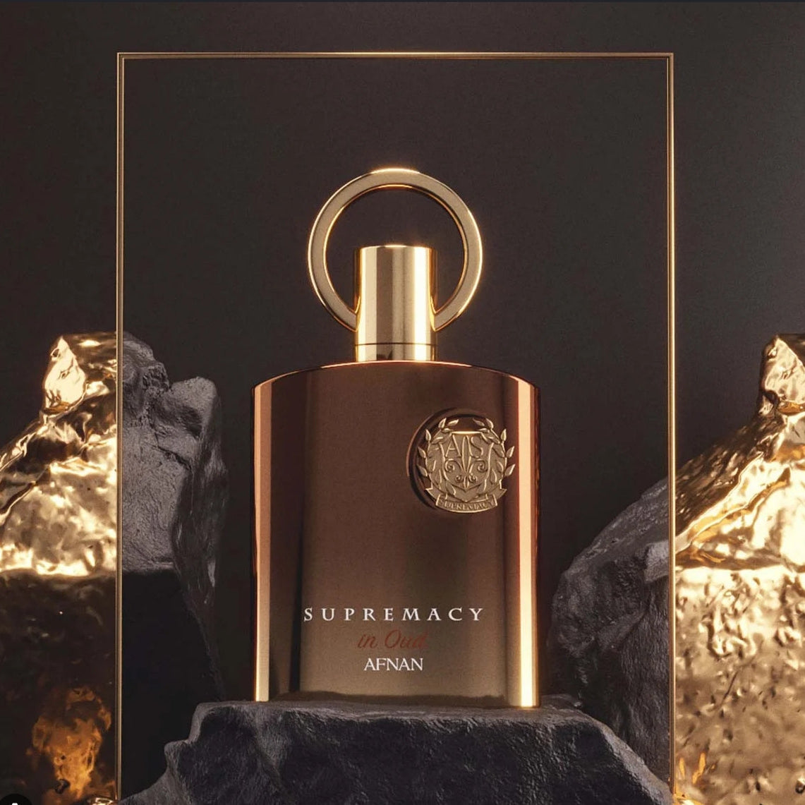 A metallic bronze and gold perfume bottle labeled 'SUPREMACY by AFNAN' displayed on a dark pedestal with textured gold rocks in the background. SHE GLOW