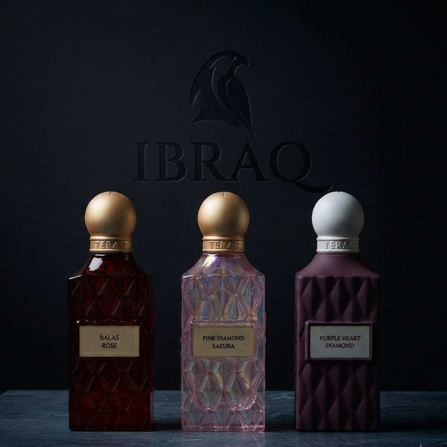 Three perfume bottles from the brand IBRAQ are lined up on a dark surface against a dark background. The bottles are faceted and contain deep red, pink, and dark purple liquids, topped with gold and white spherical caps. SHE GLOW