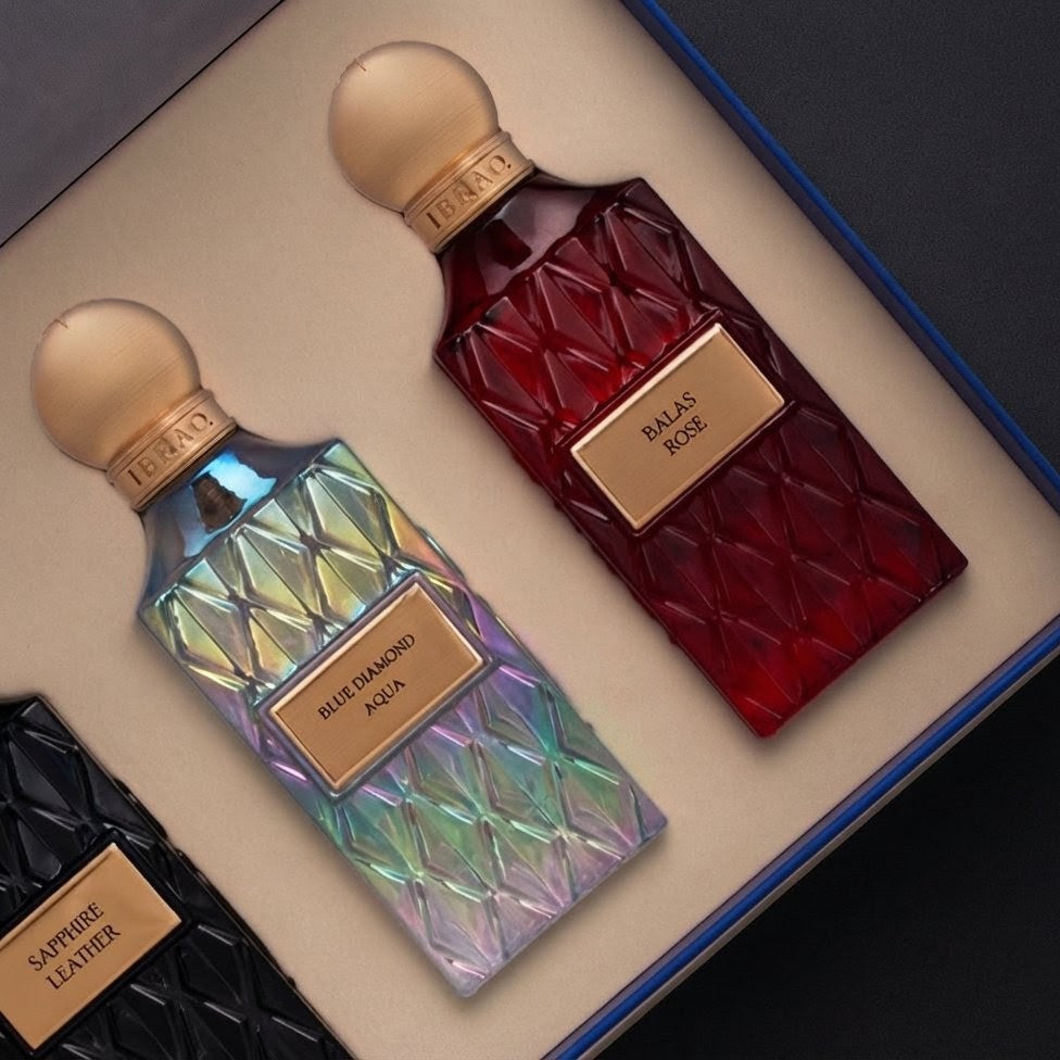 A luxury fragrance gift set in a blue box, featuring three textured glass bottles: a black 'Sapire Leather' bottle, a red 'Balas Rose' bottle, and an iridescent blue-purple 'Blue Diamond Aqua' bottle, all with gold-tone caps and labels. SHE GLOW 