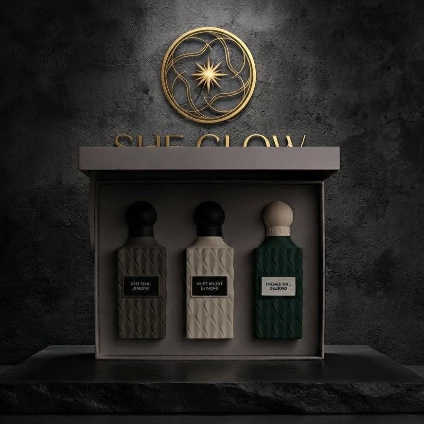 Three perfume bottles displayed inside a rectangular presentation box on a dark stone pedestal, with a textured dark gray wall behind. The bottles are textured and come in black, cream, and dark green.         SHE GLOW 