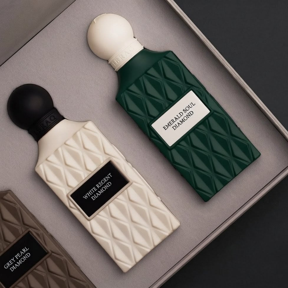 Two perfume bottles in a gray presentation box: a cream bottle labeled White Berry Diamond with a black cap, and a dark green bottle labeled Dark Alder Wood Diamond with a white cap. A partial third bottle is visible at the lower left. SHE GLOW 