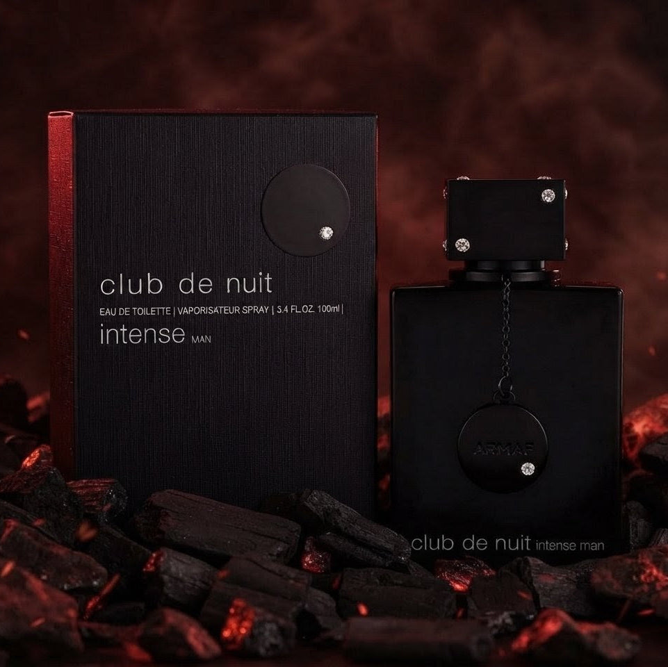 Club de Nuit Intense Man perfume bottle and its black packaging box, styled on dark charcoal briquettes against a smoky, dark red-brown background. SHE GLOW