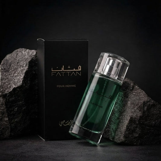 A bottle of Fattan Pour Homme fragrance with a dark green liquid in a clear glass bottle, shown next to its black packaging box with gold lettering, set against a dark background with textured rocks. SHE GLOW