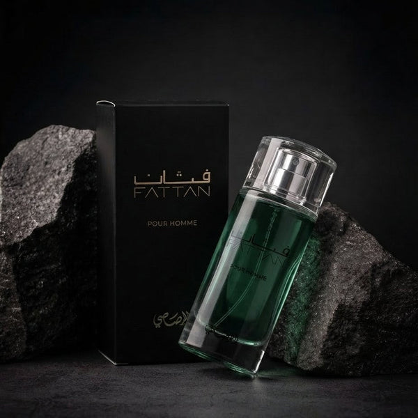 A bottle of Fattan Pour Homme fragrance with a dark green liquid in a clear glass bottle, shown next to its black packaging box with gold lettering, set against a dark background with textured rocks. SHE GLOW
