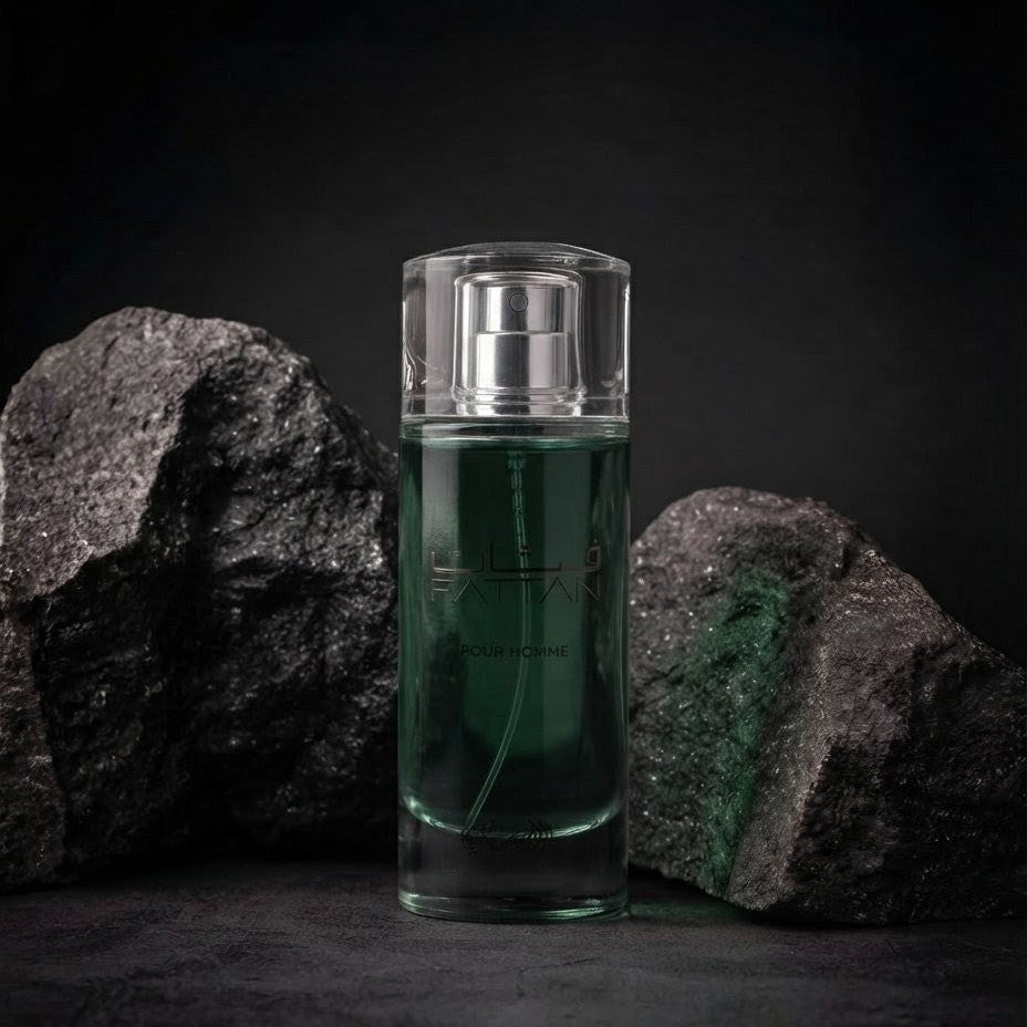 A clear rectangular perfume bottle filled with deep green liquid and topped with a silver atomizer, standing between two dark textured rocks against a black background. SHE GLOW 