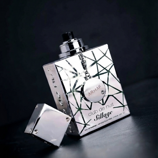 A silver metallic perfume bottle with a geometric black line pattern and a hanging round charm, shown on a dark reflective surface with the cap placed beside it. Visible text on the bottle reads 'ARMAR', 'club de nuit', and 'Sillage'. SHE GLOW