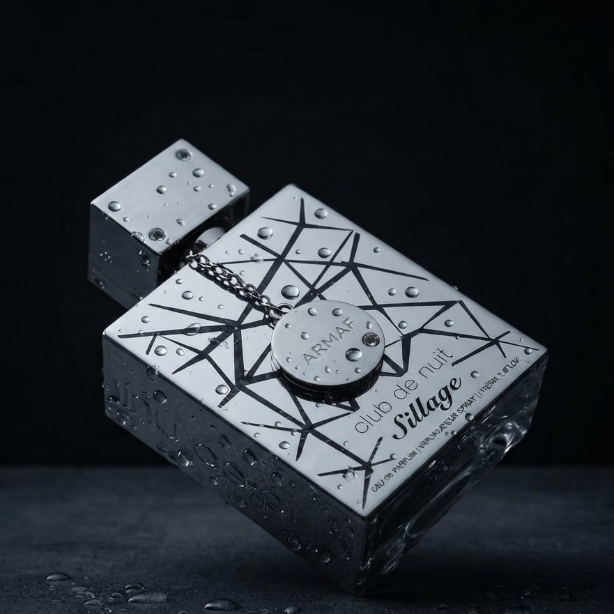 A rectangular perfume bottle with a metallic silver and black geometric pattern, covered in water droplets, floating against a dark background. The bottle features the text 'CLUB DE NUIT Village' and a circular tag reading 'PRIVE'. SHE GLOW