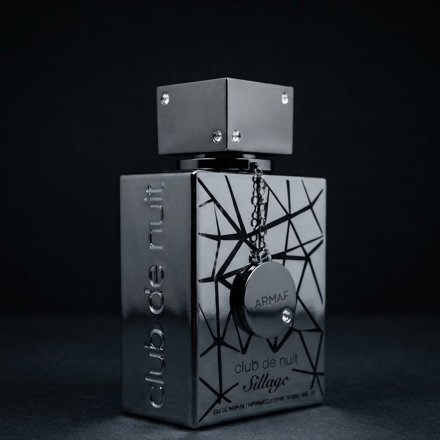 A metallic silver rectangular perfume bottle with a geometric black line pattern on the front, featuring a hanging circular tag and a matching cap, labeled 'club de nuit' and 'Sillage' with the brand 'ARMAE'. SHE GLOW