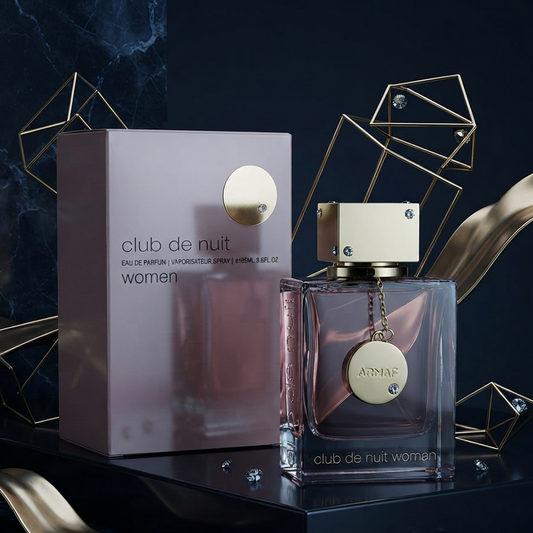 Club de Nuit Women perfume bottle and its matching box, styled on a dark reflective surface with gold geometric wireframe props and metallic ribbon accents. SHE GLOW