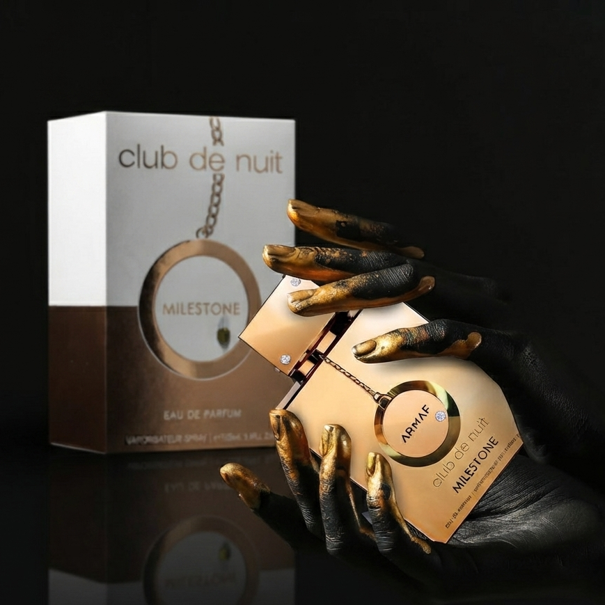 A perfume bottle and its retail box displayed on a black reflective surface. The bottle is held by a hand with long black nails and metallic gold tips, while the box stands behind it. SHE GLOW