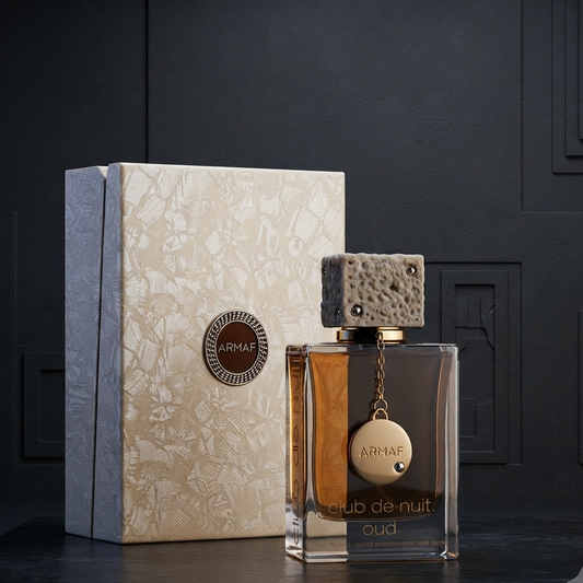 A bottle of ARMAF Oud Club de Nuit fragrance with a textured stone-like cap and gold-toned pendant, displayed next to its cream-colored patterned box on a dark surface against a dark paneled background. SHE GLOW
