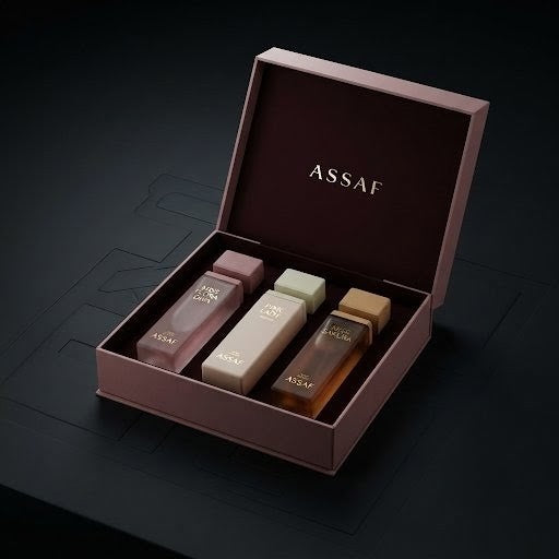 An open Assaf perfume gift box in dusty rose, containing three small fragrance bottles in pink, beige, and amber tones, set against a dark background. SHE GLOW