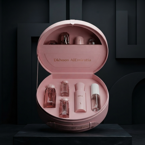 An open round pink presentation case displayed on a dark pedestal, containing several small pink and clear cosmetic bottles. The inside lid is printed with the text 'Dkhoon AlEmiratia'. SHE GLOW