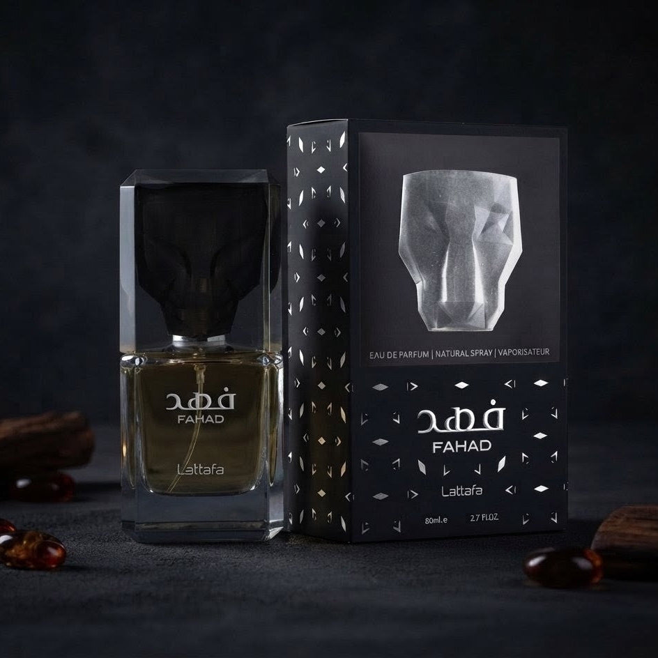 A bottle of Fahad Lattafa perfume with a black cap and clear glass, shown next to its black packaging box with gold patterns and a silver face illustration, set against a dark textured background with scattered amber beads. SHE GLOW ✨