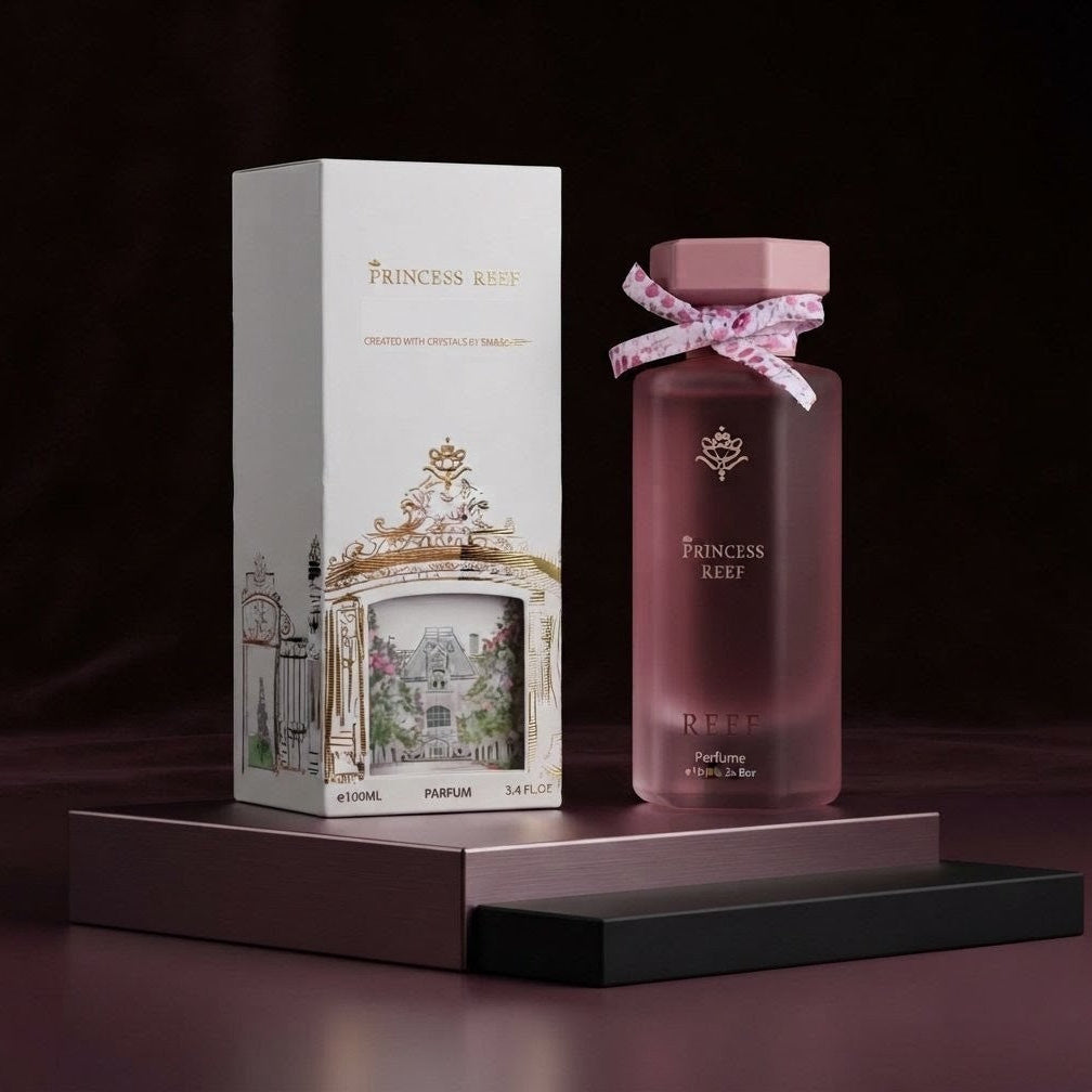 Princess Reef perfume bottle and matching box displayed on a dark pedestal. The bottle is frosted pink with a pink cap and tied with a pink patterned ribbon, and the box is white with gold detailing and a garden illustration. 
SHE GLOW ✨