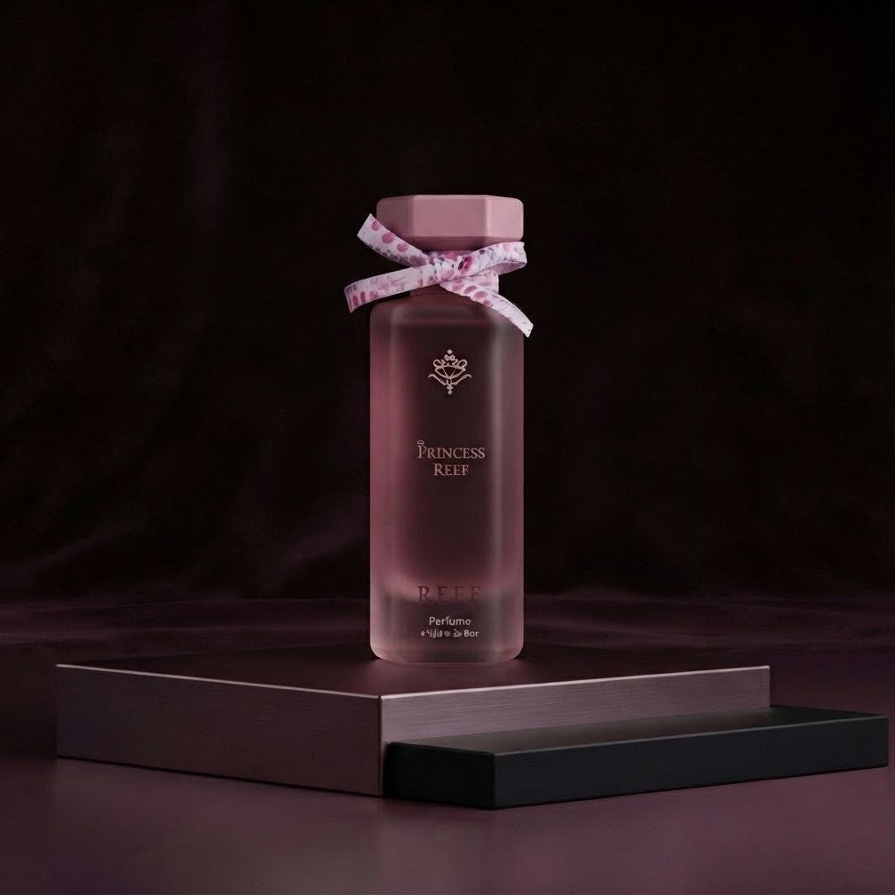 A frosted glass perfume bottle with a dusty rose pink cap and a matching pink ribbon bow tied around the neck, standing on a dark metallic display block against a dark brown background. The bottle features gold lettering that reads 'PRINCESS RIFE' and 'EAU DE PARFUM'.
SHE GLOW ✨