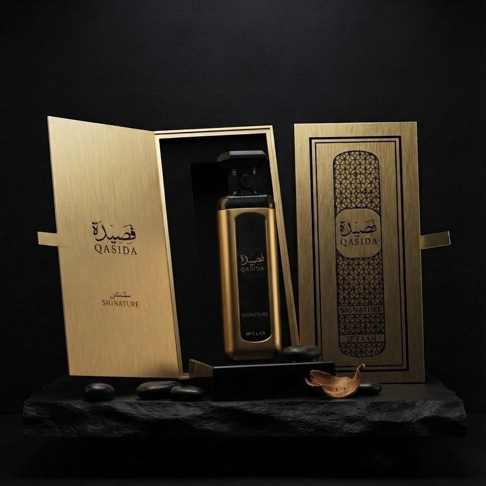 A luxury perfume bottle in a brushed gold finish with black detailing, presented in an open matching gold box next to its closed lid on a dark stone base. The brand name 'Qasida' is visible in Arabic and English on the packaging.SHE GLOW ✨