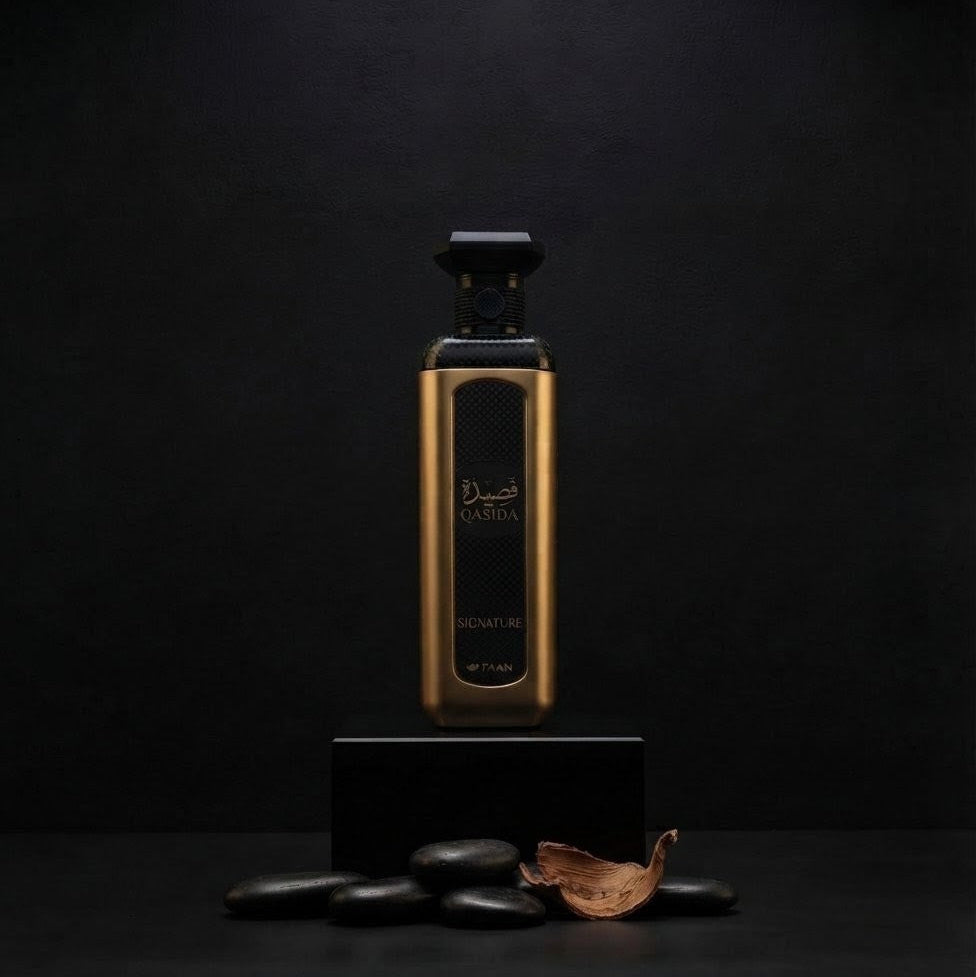 A tall rectangular perfume bottle with a metallic gold body and black cap, displayed on a black pedestal against a dark background. Arabic text is visible on the bottle, and a few dark stones and a dried leaf sit at the base. SHE GLOW ✨