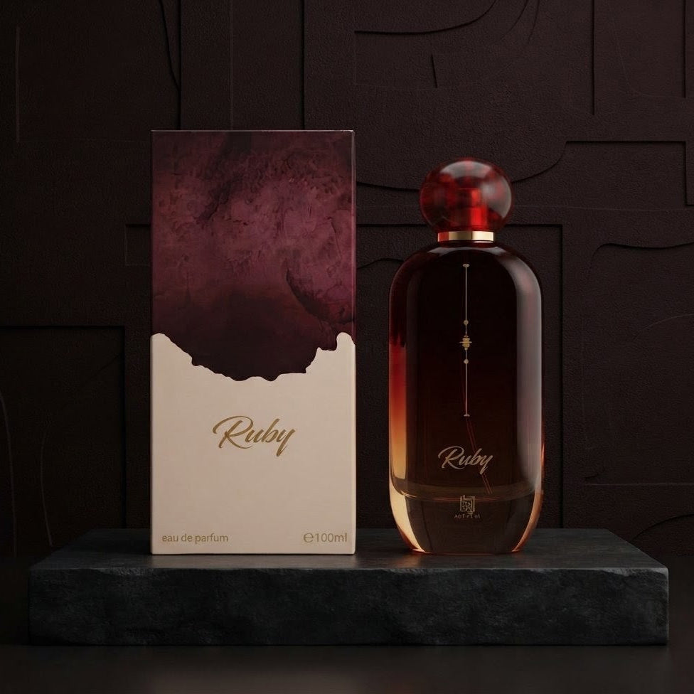 A bottle of Ruby Eau de Parfum with a dark amber glass body and a round red cap, displayed next to its matching box on a dark stone pedestal against a dark textured background.SHE GLOW ✨
