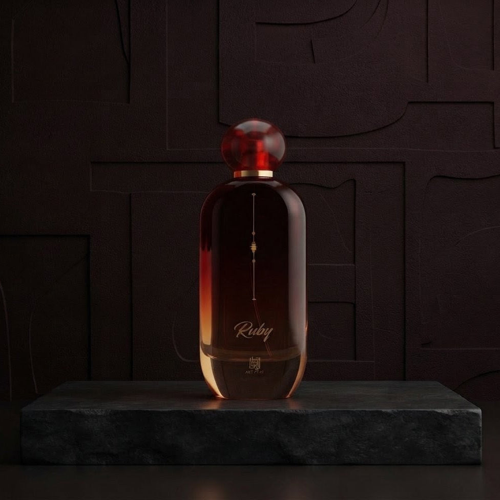 A dark amber glass perfume bottle with a spherical red cap, standing on a dark stone pedestal against a dark textured background. The bottle features the brand name 'Rubiq' in gold script.SHE GLOW ✨