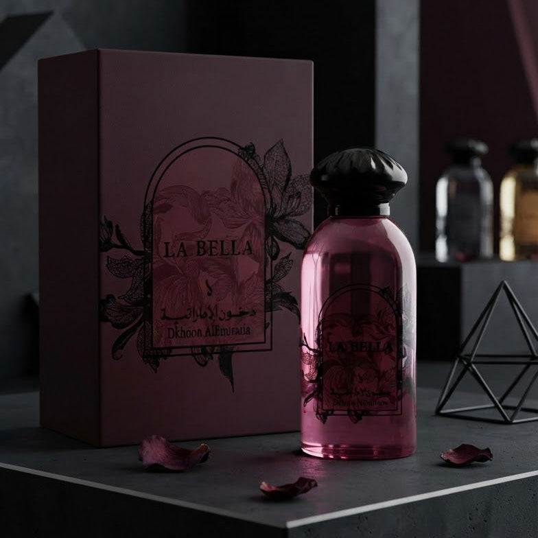 A pink glass perfume bottle with a black cap stands next to a matching dark mauve box labeled LA BELLA, styled on a dark geometric platform with scattered rose petals. SHE GLOW 