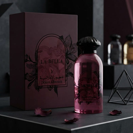A pink glass perfume bottle with a black cap stands next to a matching dark mauve box labeled LA BELLA, styled on a dark geometric platform with scattered rose petals. SHE GLOW 