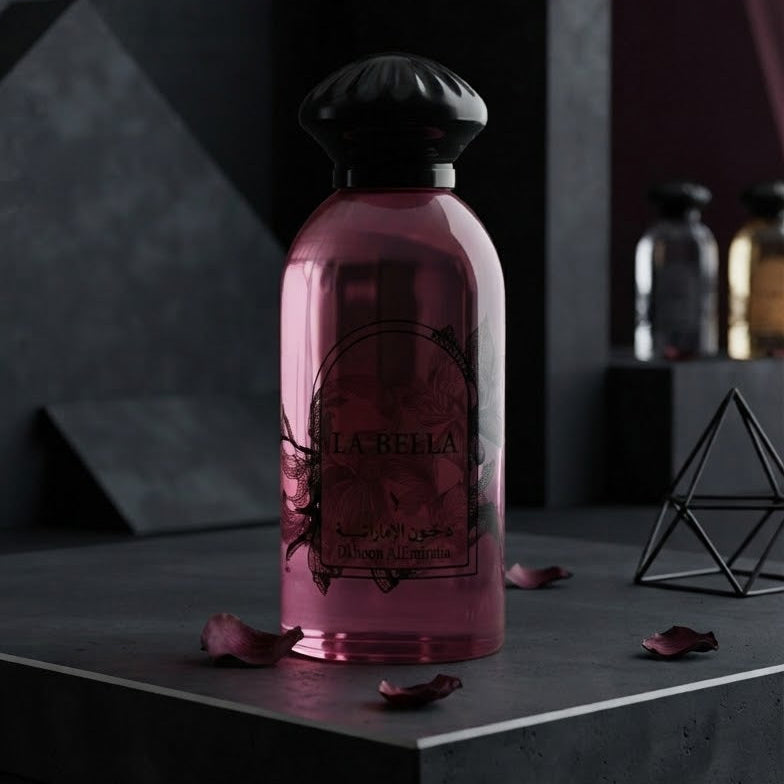 A pink glass perfume bottle with a black cap, labeled 'L'Abri', displayed on a dark gray pedestal with scattered rose petals and dark geometric background props. SHE GLOW 