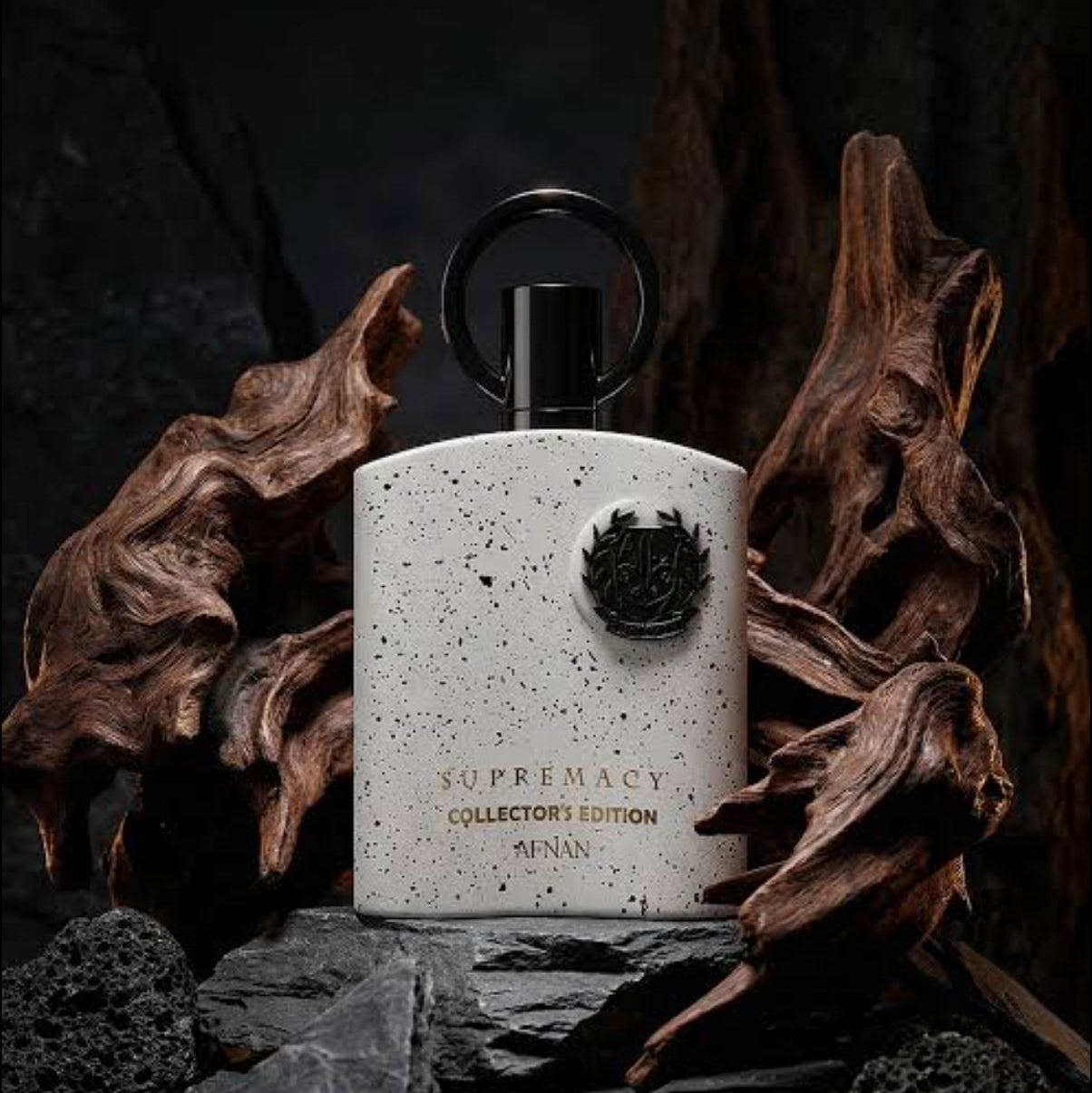 A white speckled perfume bottle with a black cap and ring handle, labeled 'Supremacy Collector's Edition AFAN', styled on dark rocks and driftwood against a dark background. SHE GLOW